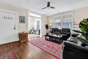A living room with a dark leather sofa, a coffee table, and a red patterned rug. There is a ceiling fan, a wooden cabinet, and a large plant.