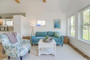 Un salon avec un canapé bleu, un fauteuil blanc, une table basse blanche et une fenêtre donnant sur un paysage verdoyant.
