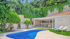 A swimming pool with a curved edge, surrounded by a patio area with lounge chairs and a covered seating area. - Villa Banus (Marbella)