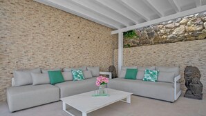 A modern living room with a brick wall, a white ceiling, and a stone wall. There are two grey sofas with green pillows and a white coffee table with a vase of flowers. - Villa Banus (Marbella)