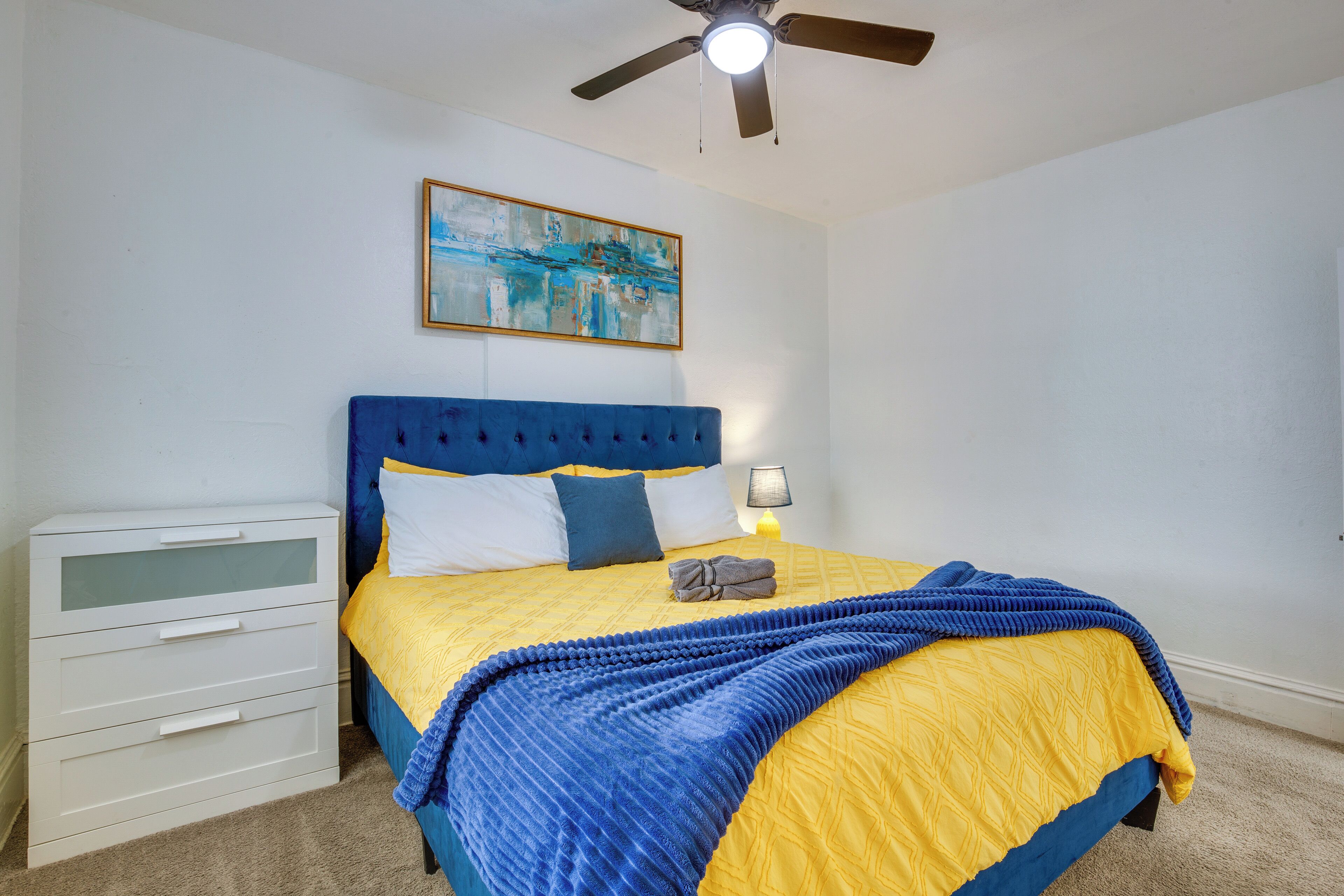 A bedroom with a bed, a nightstand, a ceiling fan, and a painting on the wall.