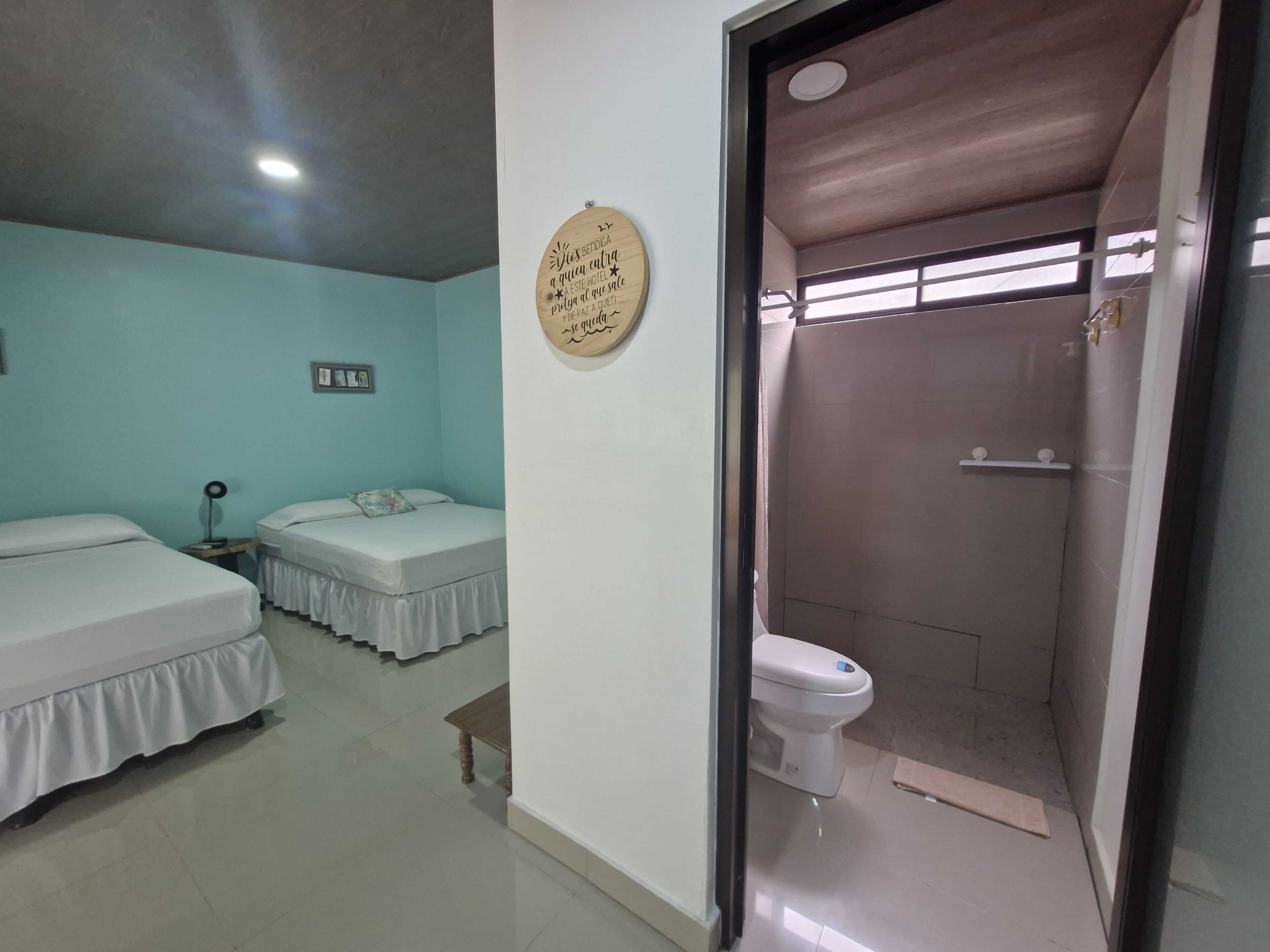 A room with two beds, a small table, a wall-mounted light, and a shower area with a toilet.