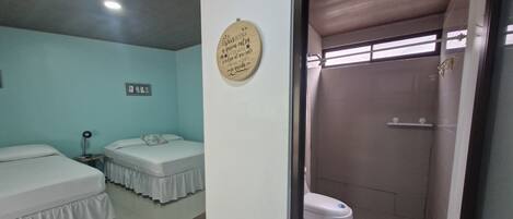 A room with two beds, a small table, a wall-mounted light, and a shower area with a toilet.