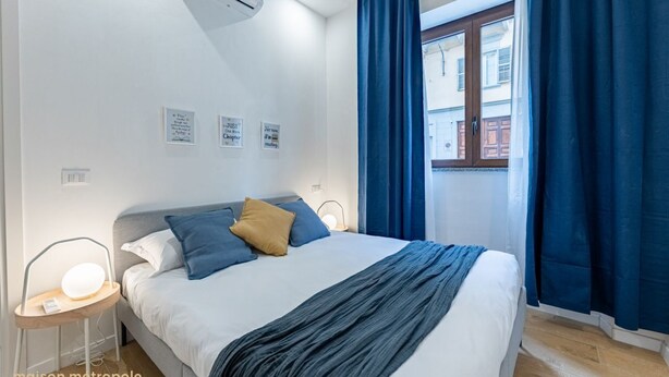 Une chambre à coucher avec un lit, une table de chevet avec une lampe, une fenêtre avec des rideaux et des œuvres d’art murales.