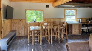 A rustic cabin interior with wooden walls and ceiling, a dining area with mismatched wooden chairs, a kitchen with a wooden counter, and a large window overlooking greenery.