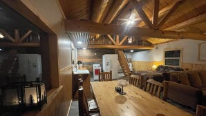 A cozy cabin interior with a wooden dining table, chairs, a sofa, and a kitchen area with a refrigerator and stove.