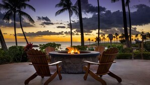A fire pit with chairs around it, overlooking a beach with palm trees and a sunset.