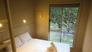 A neatly made bed with white linens and a folded towel, a bedside table, a window with a view of greenery, and a wall-mounted light fixture. - FDE 30 - FAMILY DELUXE 3 BEDROOM CABIN (Cape Otway)