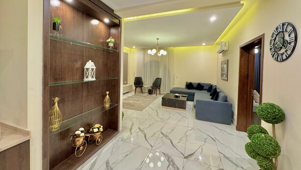 A modern living room with a marble floor, a dark wood shelving unit with decorative items, a blue sofa, and a large wall clock.