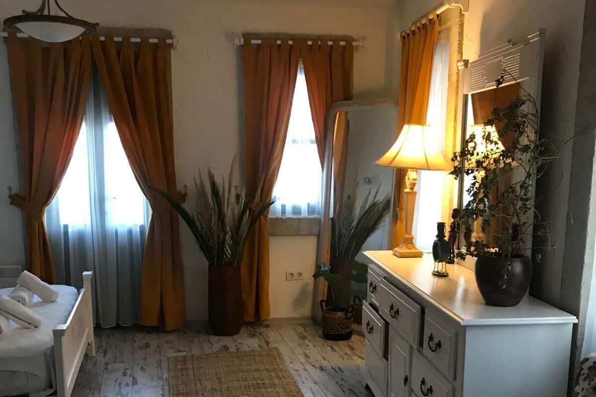 Une pièce comprenant un lit, une commode, une chaise et un miroir. Il y a deux fenêtres avec des rideaux, un tapis sur le sol et une plante sur la commode.