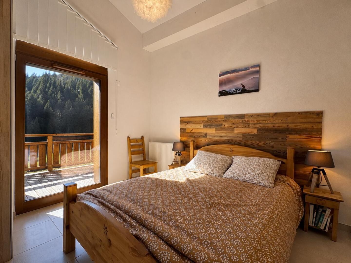 A bedroom with a wooden headboard, a bed with patterned bedding, a nightstand with a lamp, and a view of a forest through a large window.