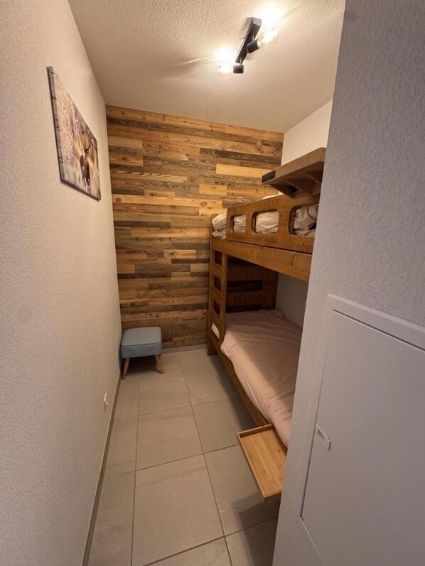 A small, cozy room with a wooden wall panel, a bunk bed, a small stool, and a door.