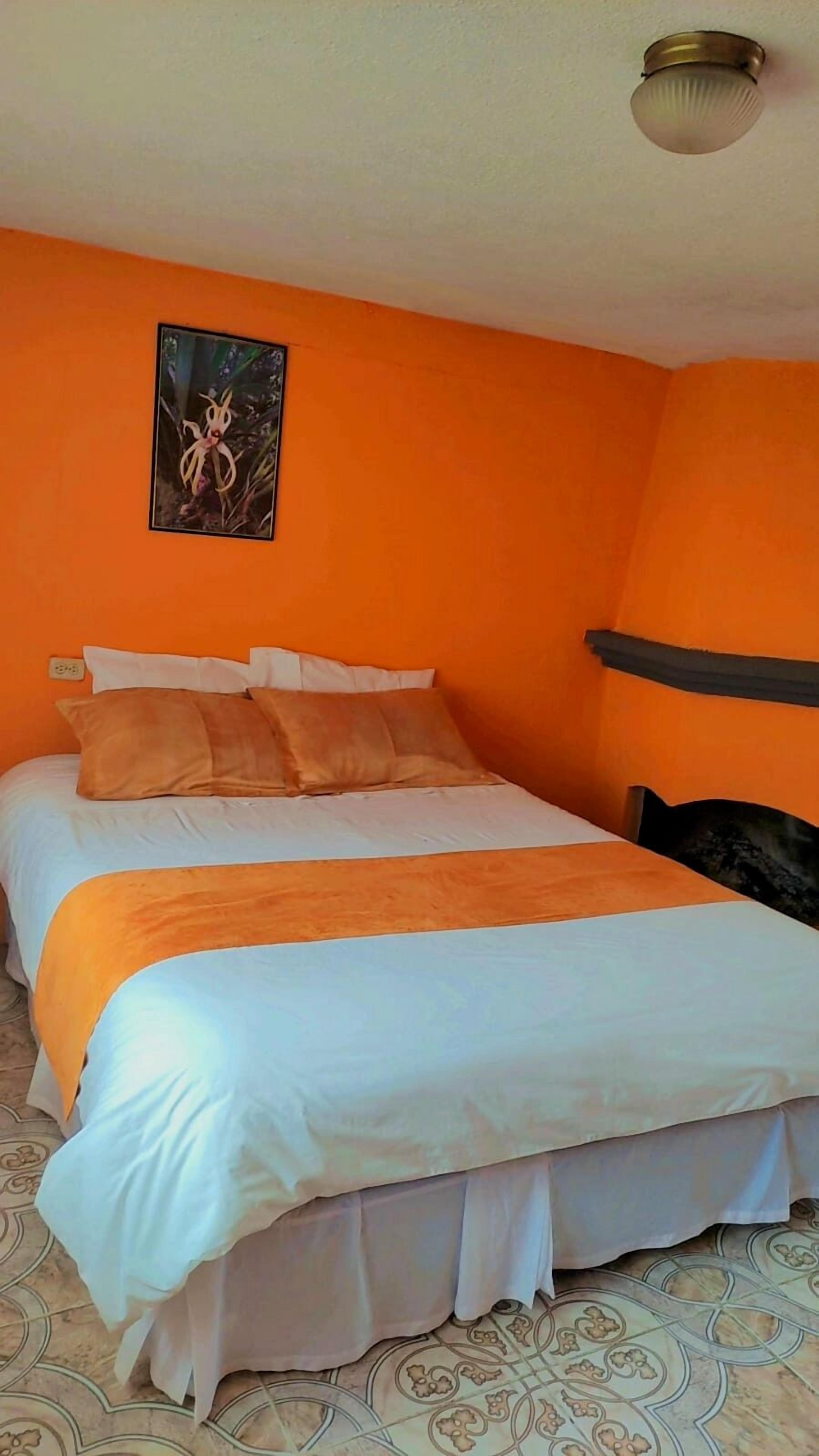A single bed with white and orange bedding, a nightstand, and a wall-mounted light fixture in a room with orange walls and patterned floor tiles.