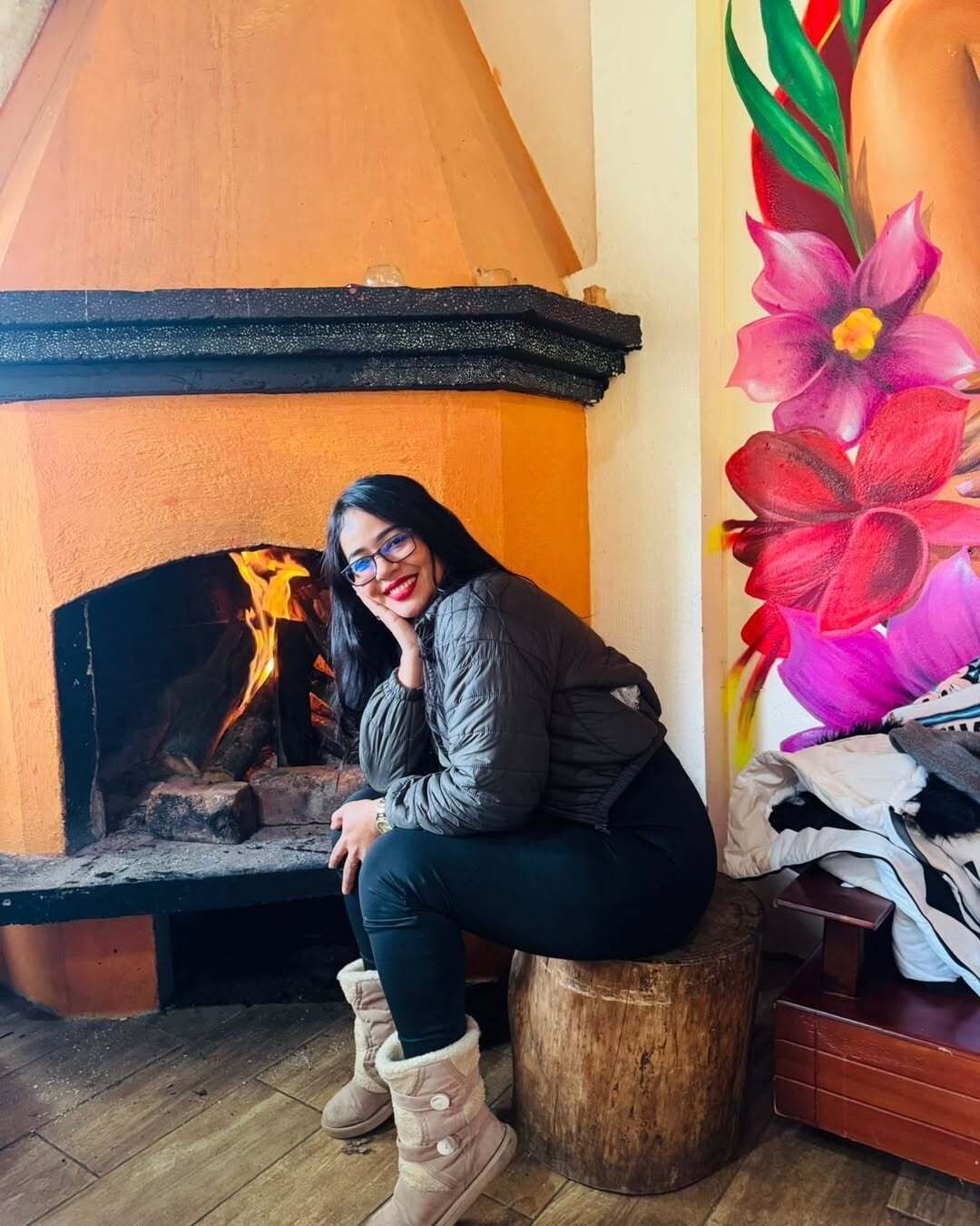 Una persona sentada junto a una chimenea, con un cuadro de flores en la pared.