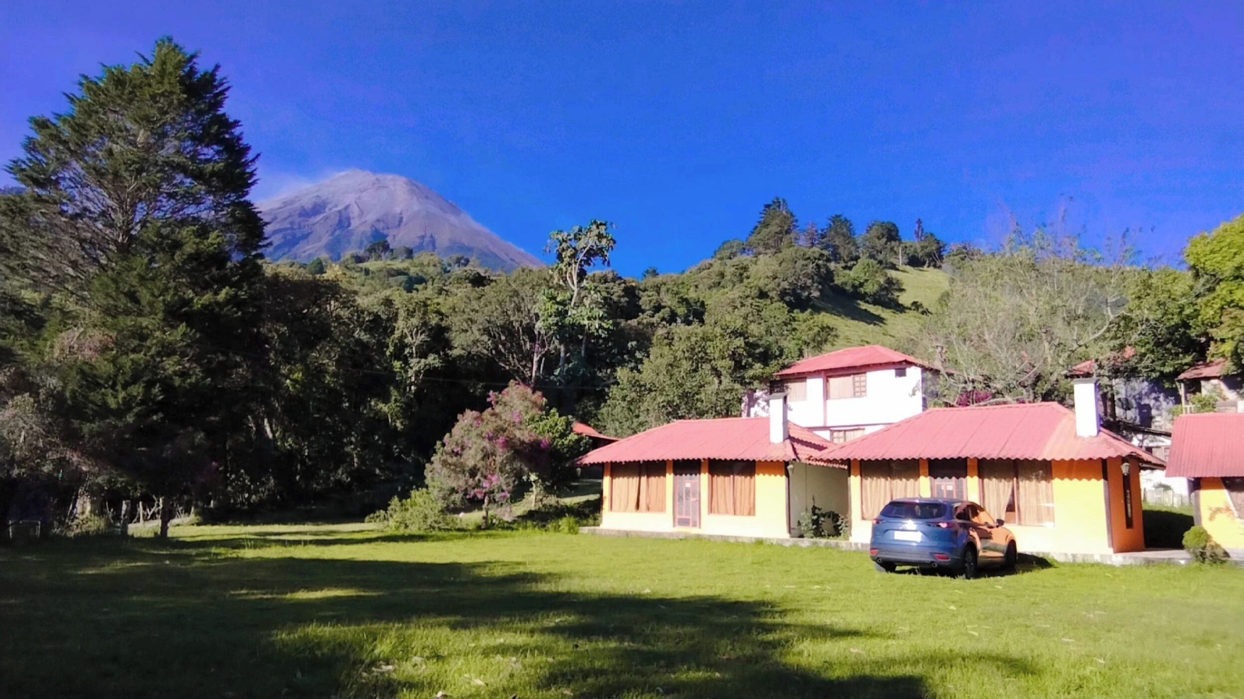Una casa con techo rojo, un coche estacionado adelante y una montaña al fondo.