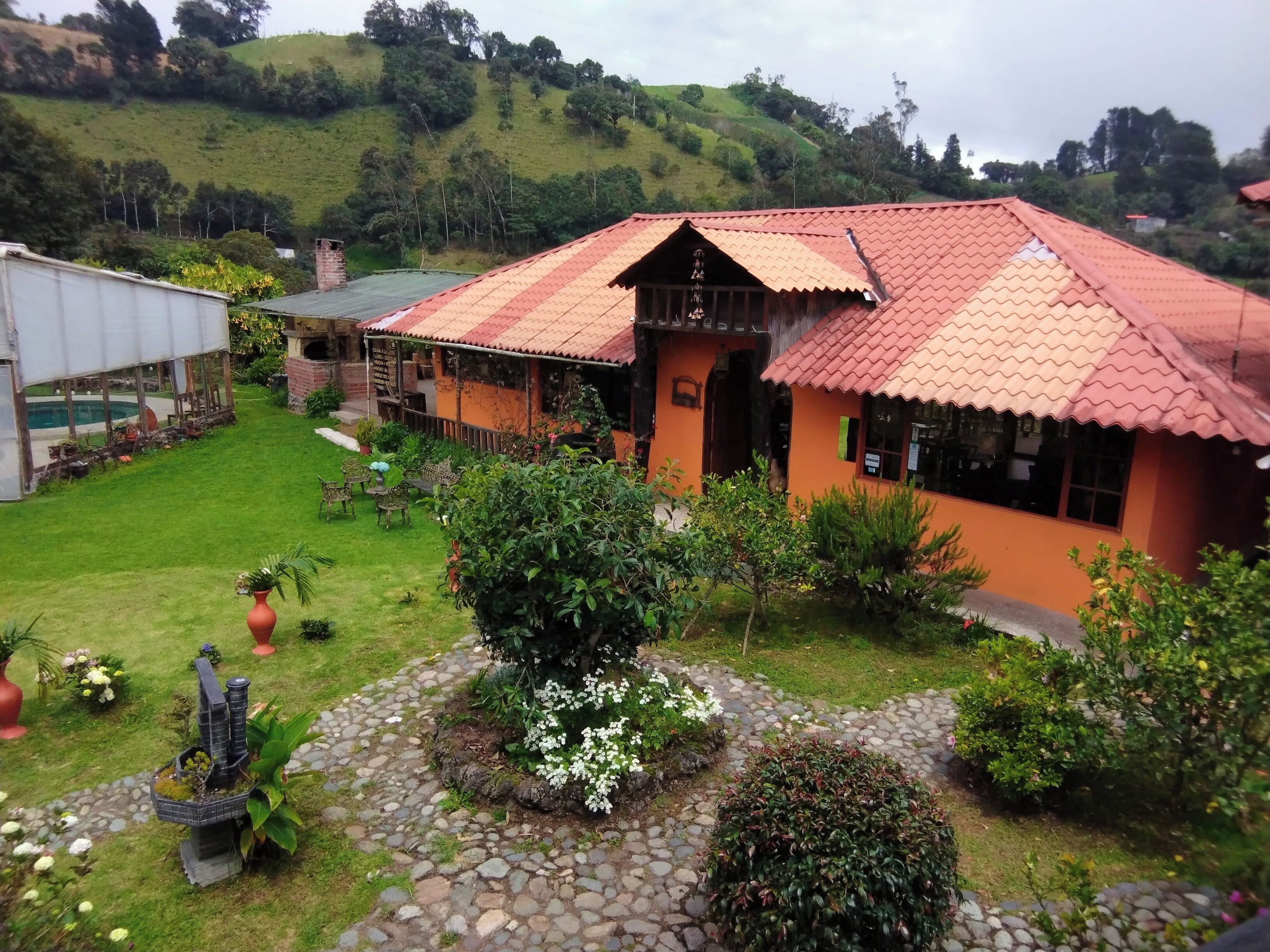 A building with a red-tiled roof, surrounded by a garden with a stone pathway, and a hilly landscape in the background.