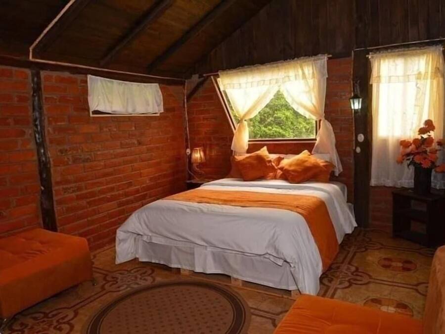 A rustic room with a brick wall, a bed with white and orange bedding, a small orange ottoman, and a window with curtains.