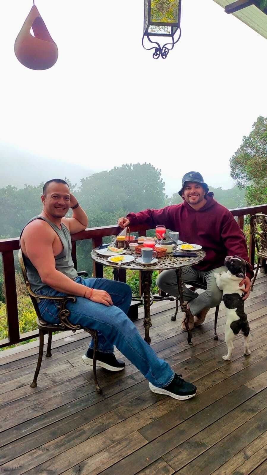 Two people sitting at a round table with food and drinks on a wooden deck with a hanging lantern.