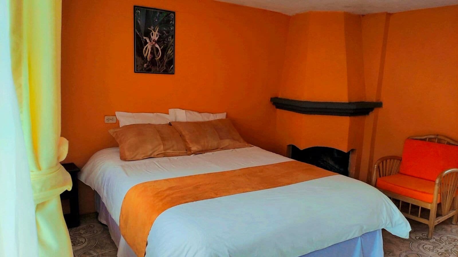 Ein Schlafzimmer mit einer orangefarbenen Wand, einem Bett mit weißer und orangefarbener Bettwäsche, einem Sessel mit rotem Kissen und einem Blumenbild.