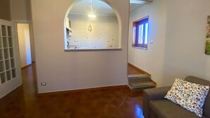 Un amplio salón con suelo de madera, un sofá gris y un cojín estampado. Se ve una cocina a través de un arco, y una escalera conduce a otro nivel.