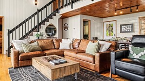 A spacious living room with a large brown leather sectional sofa, a wooden coffee table, and a black leather armchair. A staircase with black railings leads to an upper level. - 6 Bedroom VIEW next to Dollywood FLASH SALE today (Sevierville)