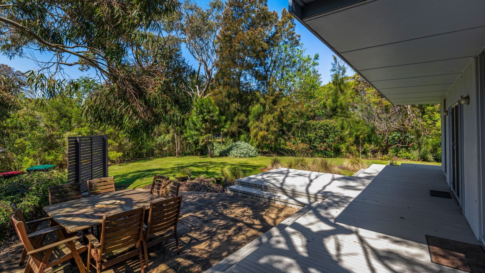 A covered patio with a wooden table and chairs, surrounded by a lush garden and tall trees.