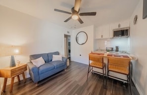 A compact living space with a sofa, a dining table with chairs, a ceiling fan, and a kitchen area.