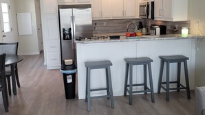 Cocina moderna con armarios blancos, un refrigerador de acero inoxidable y una encimera de mármol. Hay tres taburetes de bar y un cubo de basura.