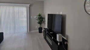 Una sala de estar moderna con un televisor de pantalla plana, una planta en maceta y un amplio ventanal con persianas.