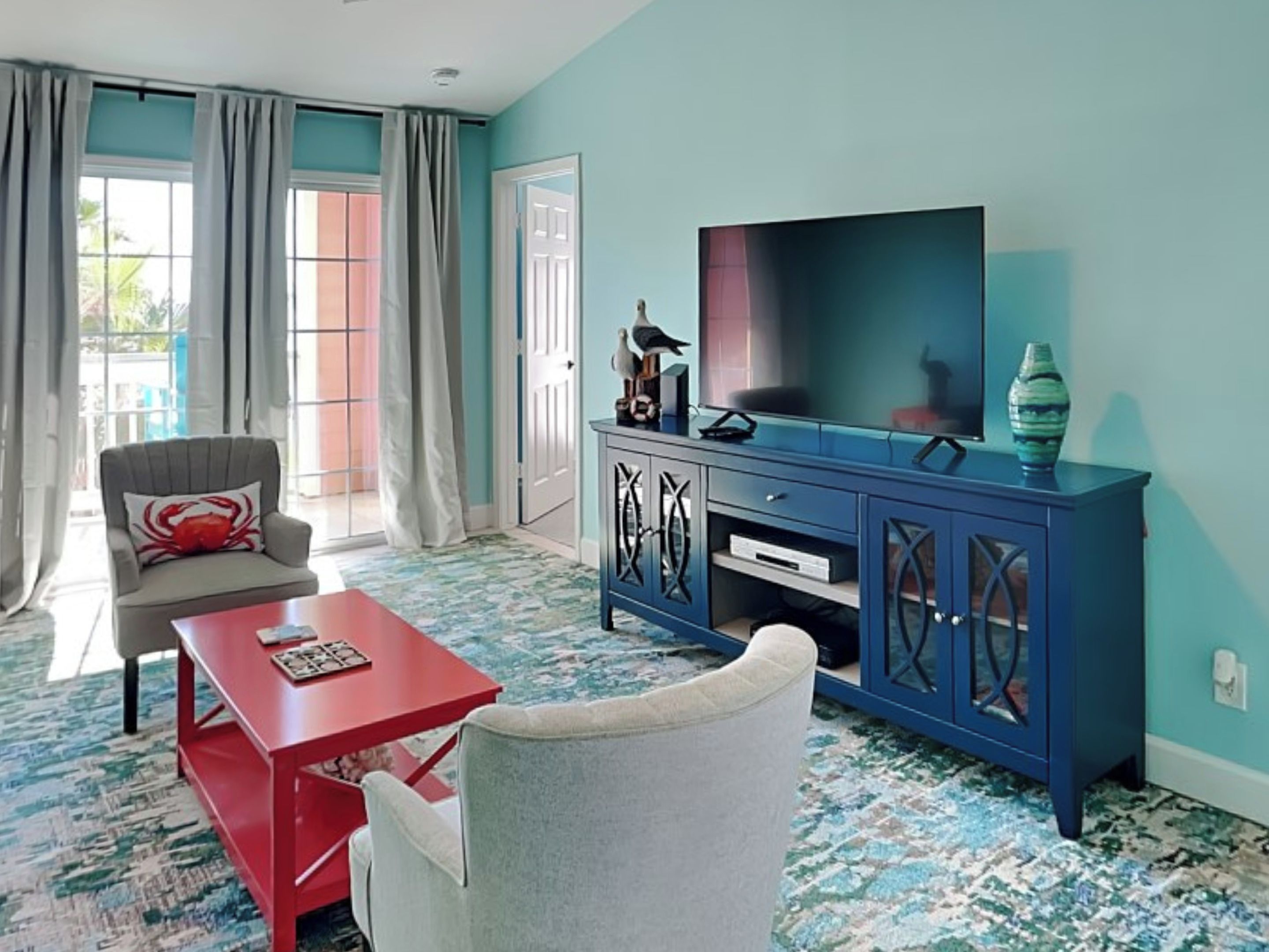 Un salon avec une table basse rouge, un fauteuil gris, un meuble bleu contenant un téléviseur et un tapis à motifs.