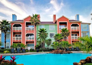 A hotel with colorful buildings, a swimming pool, and palm trees. - Beach and Pool View by Ryson Vacation Rentals (Galveston)