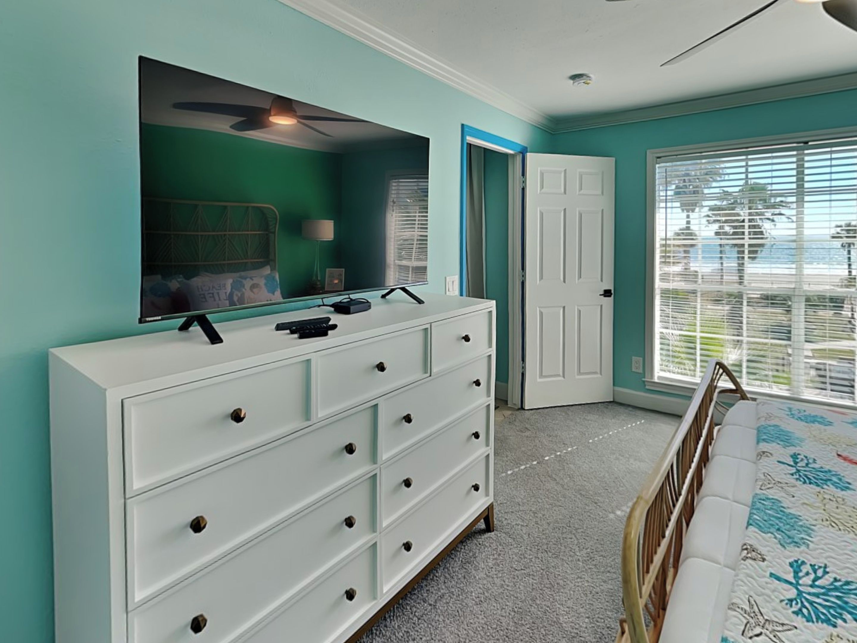 Une chambre avec une commode blanche, un lit recouvert d’une couvre-lit à motifs floraux, un ventilateur de plafond et une fenêtre avec des stores.