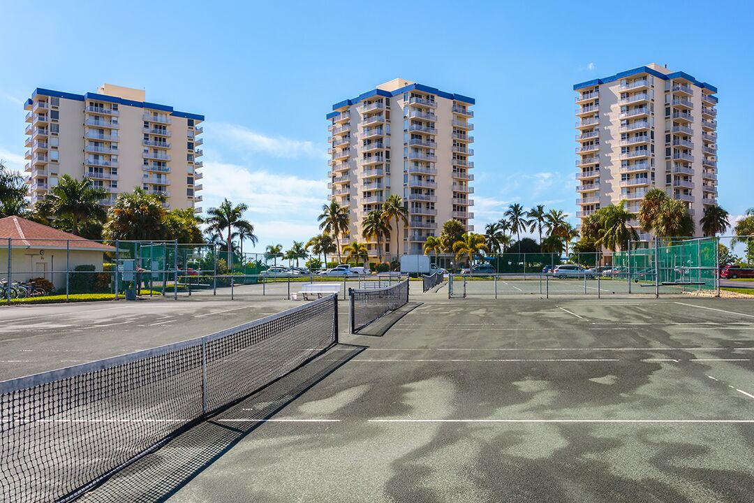 A tennis court with a net, surrounded by palm trees and tall buildings.
