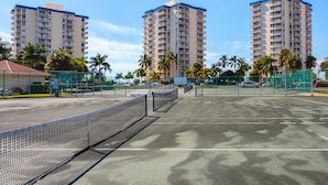A tennis court with a net, surrounded by palm trees and tall buildings.