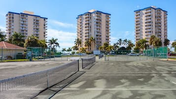 A tennis court with a net, surrounded by palm trees and tall buildings.