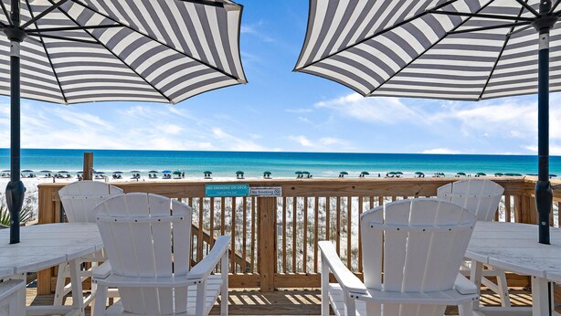 A wooden deck with white chairs and striped umbrellas overlooking a beach. - Capri on the Gulf 105 (Destin)