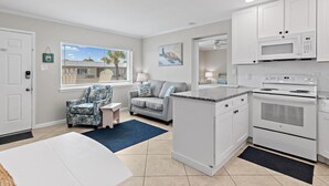 A modern kitchen with white cabinets, a built-in microwave, and a double oven. There is a gray sofa and a patterned armchair in the adjacent living area. - Capri on the Gulf 105 (Destin)