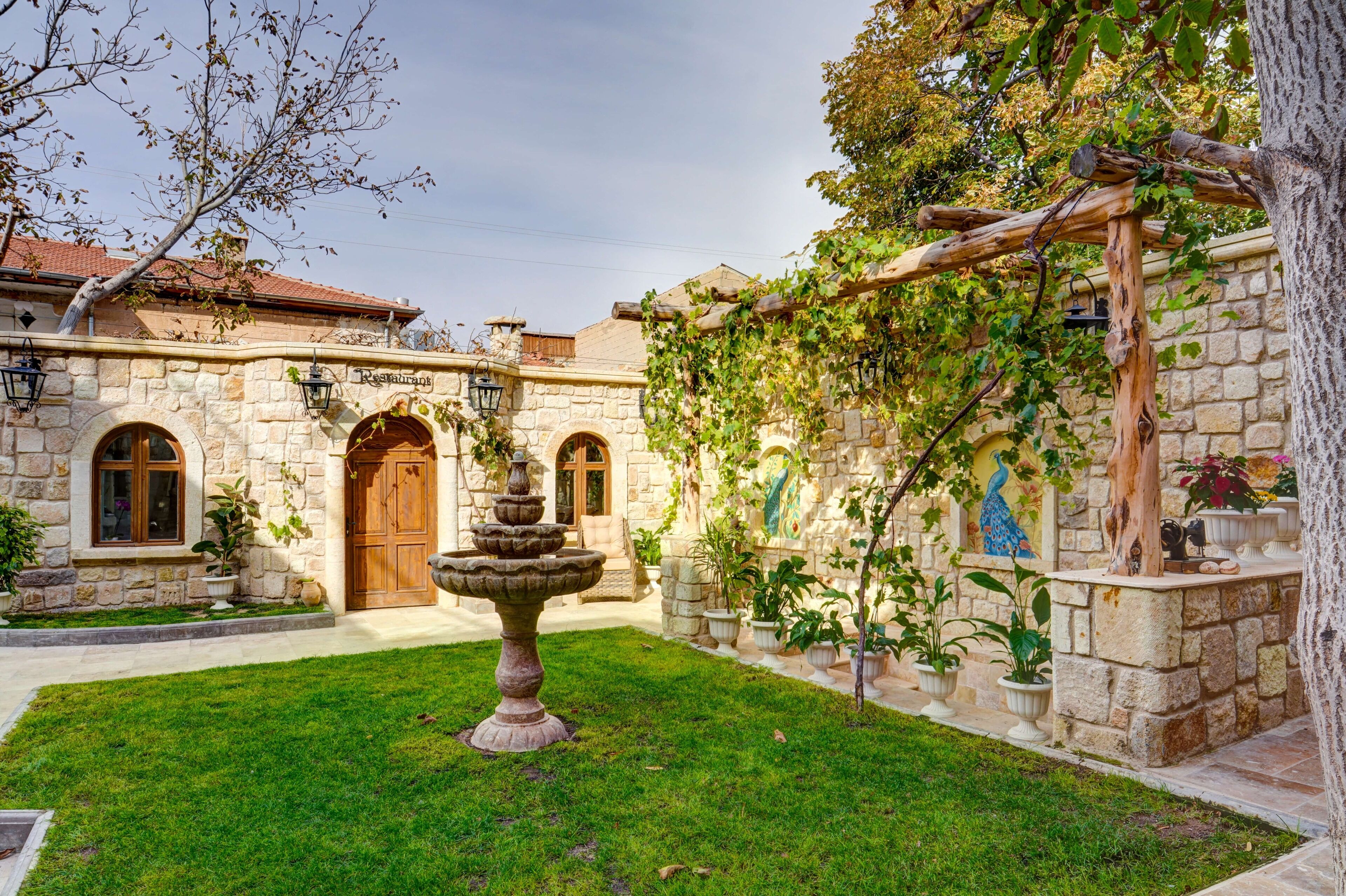 Un patio con una fuente, muros de piedra y una pérgola cubierta de plantas trepadoras.