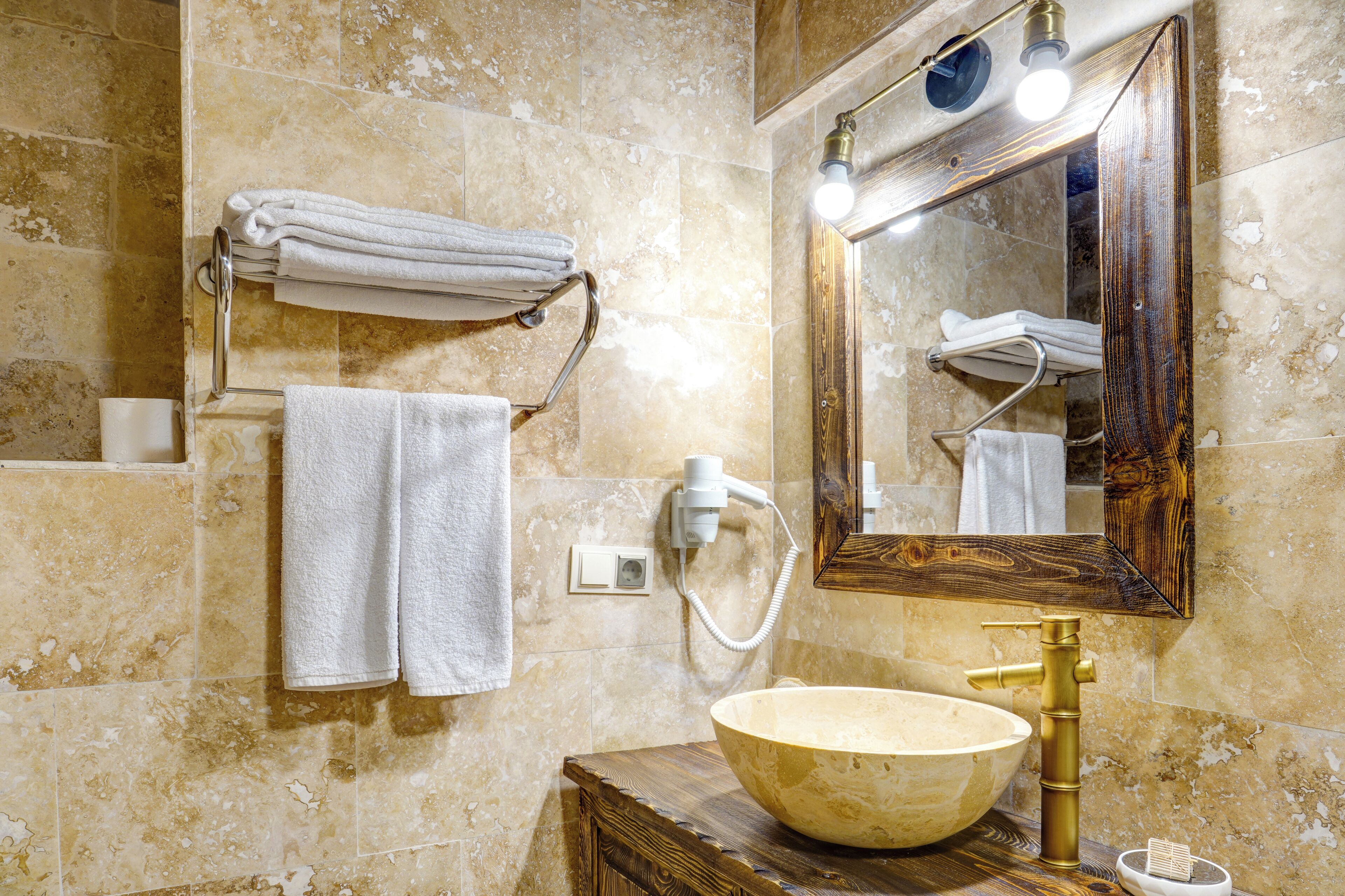 A bathroom with a large bowl sink, a wooden vanity, a towel rack with folded towels, a mirror with lights, and a hairdryer on the wall.