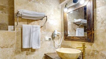 A bathroom with a large bowl sink, a wooden vanity, a towel rack with folded towels, a mirror with lights, and a hairdryer on the wall.