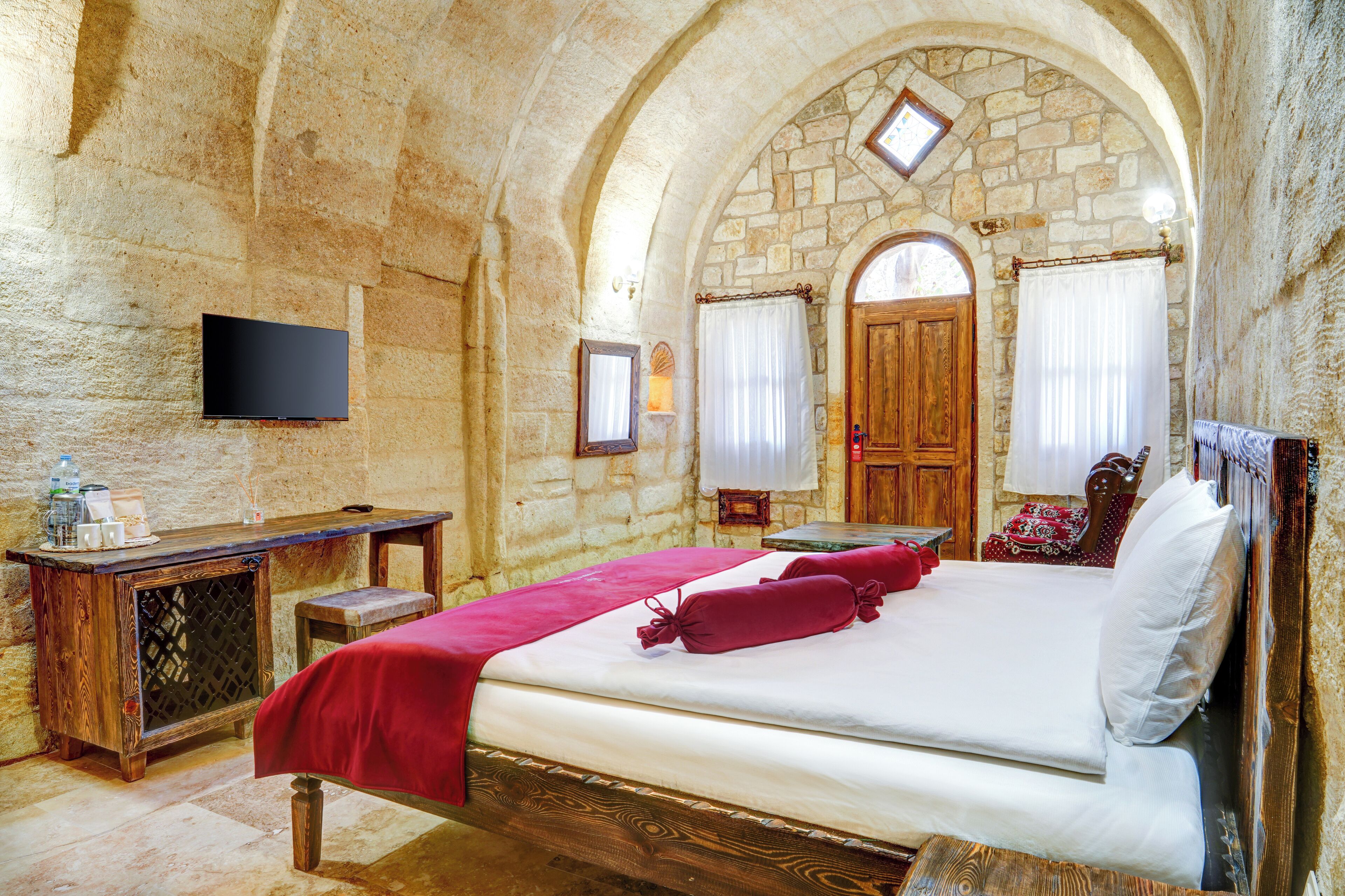 A stone-walled bedroom with a bed, a wooden desk, a television, and a small table.