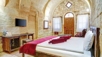 A stone-walled bedroom with a bed, a wooden desk, a television, and a small table.