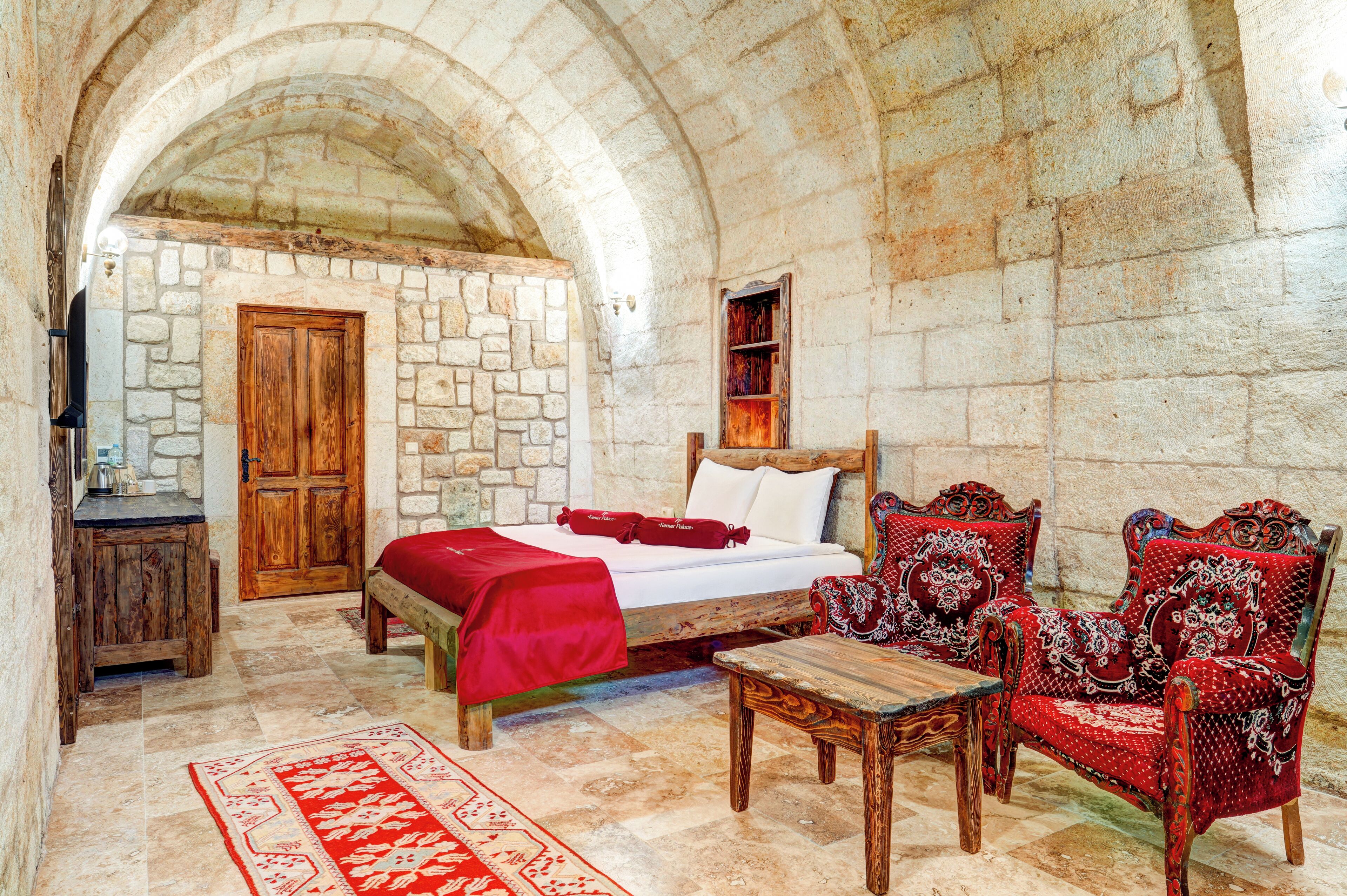 A stone-walled room with a bed, wooden furniture, and a red rug.