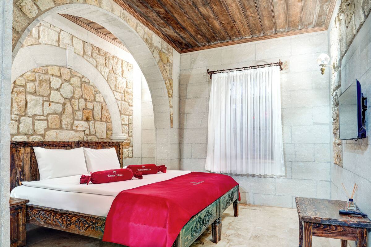 Un dormitorio con pared de piedra, techo de madera, cama con sábanas blancas y manta roja, una mesita con lámpara y ventana con cortina blanca.