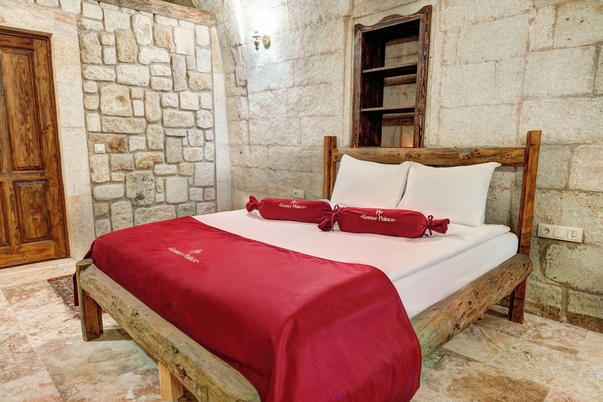 Una cama con colcha y almohadas rojas, una pared de piedra, una puerta de madera y un pequeño estante de madera.