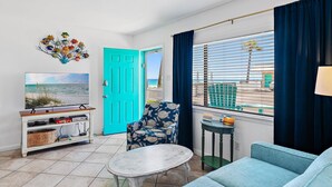 A living room with a TV, a blue chair, a small table, and a view of the beach through the window. - Capri by the Gulf 106 (Destin)