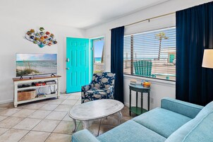 Una sala de estar con televisor, un sillón azul, una mesita y vista al mar a través de la ventana.