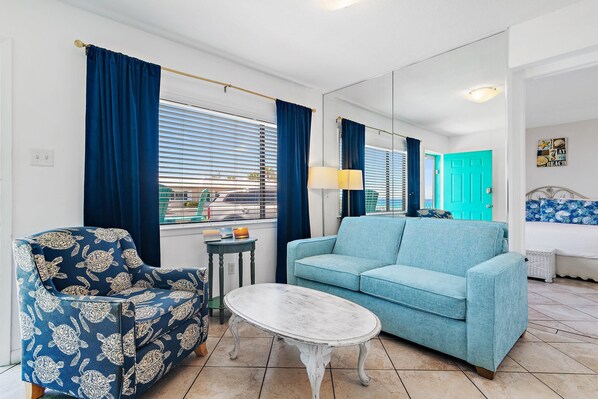 Una sala de estar con un sofá, un sillón estampado, una mesita y una ventana con cortinas.