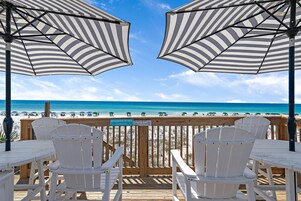 Terraza de madera con sillas blancas y sombrillas a rayas con vista a la playa.