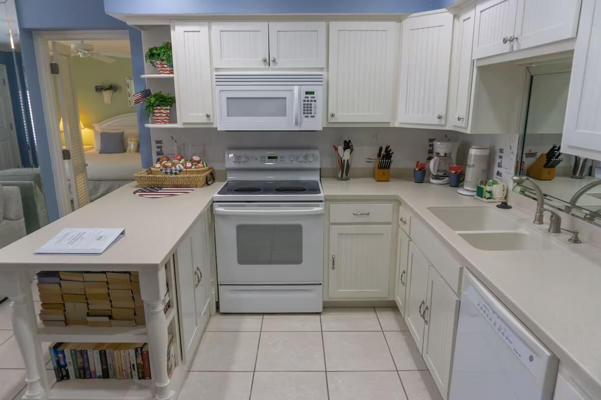 A kitchen with white cabinets, a microwave, an oven, a sink, and a dishwasher. There is a countertop with a basket of fruits and a book.